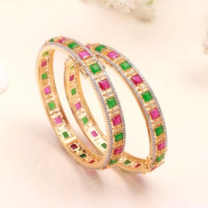 Puede incluir: Dos brazaletes dorados con bisagra que presentan piedras preciosas cuadradas alternadas en verde y rosa. Los bordes exteriores de los brazaletes están bordeados con pequeñas piedras claras y brillantes engastadas en un marco metálico.