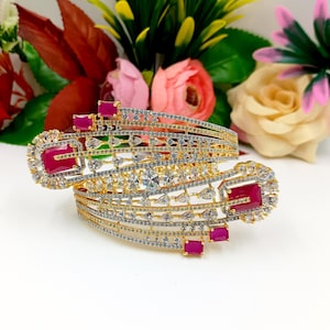 May include: Two gold-toned bangles with intricate designs. The bangles are adorned with clear and ruby-colored gemstones. The design includes rectangular and square-shaped ruby stones and clear, sparkling stones. The background features colorful flowers.