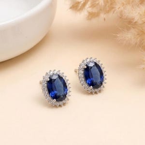Könnte beinhalten: Ein Paar Ohrstecker mit oval geschliffenen, tiefblauen Mittelsteinen, umgeben von einem Kranz aus kleinen, klaren, rund geschliffenen weißen Steinen in einer Metallfassung. Die Ohrringe verfügen über einen sicheren Steckverschluss.