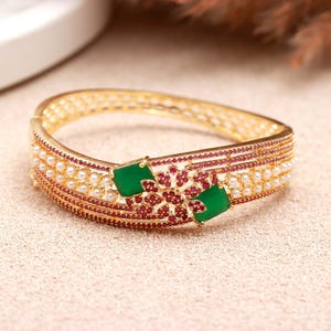 Puede incluir: Una pulsera rígida dorada con bisagra que presenta dos piedras verdes cuadradas, un motivo floral central con pequeñas piedras rojas y filas de diminutas perlas blancas. La banda metálica está decorada con patrones de pequeñas piedras rojas.