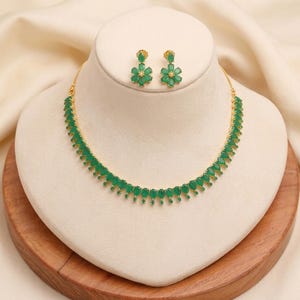 May include: A gold-toned necklace and matching earrings set. The necklace and earrings feature emerald green teardrop-shaped stones. The earrings are flower-shaped. The jewelry is displayed on a white bust against a cream-colored background.