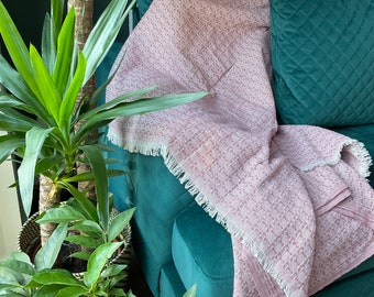 Puede incluir: Una manta de tiro rosa con flecos blancos, drapeada sobre un sofá de terciopelo verde azulado. La manta tiene un sutil patrón geométrico.