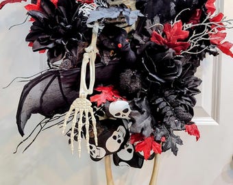 May include: A decorative wreath featuring a central skeleton figure wearing a black hat, surrounded by black and red artificial flowers, dark foliage, bat wings, and a ribbon patterned with white skulls on a black background, hanging vertically.