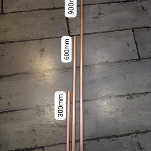 May include: Three cylindrical copper pipes arranged vertically, labelled with their respective lengths of 300mm, 600mm, and 900mm. The metallic pipes have a smooth, polished finish and are aligned side by side, showing a consistent diameter across all three pieces.