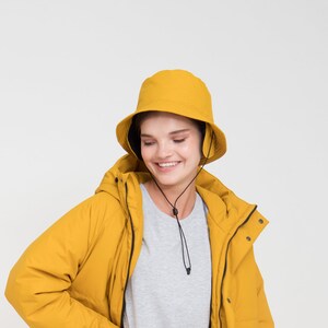 May include: A mustard yellow hooded jacket and matching bucket hat. The jacket has a zip closure and pockets. The outfit includes a gray t-shirt, gray jeans, and white boots. The person is smiling.
