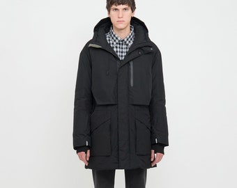 Pode incluir: Um parka preto com capuz e vários bolsos, usado sobre uma camisa xadrez e jeans escuros. O parka tem fecho de correr e um design aerodinâmico. O modelo usa tênis pretos com solas marrons. O fundo é branco liso.
