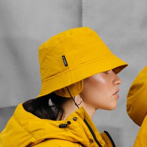 May include: A person wearing a bright yellow bucket hat and matching jacket. The hat has a small black tag on the side. The jacket has a zipper and a hood. The background is a light gray.