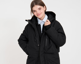 May include: A long black winter coat with a hood, multiple pockets, and a zipper closure. The coat is worn over a collared shirt and a dark sweater. The model is smiling and standing in front of a white background.