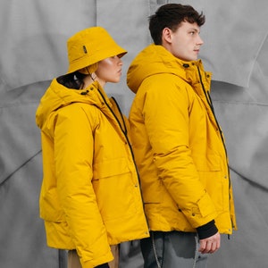 May include: Two people wearing matching yellow hooded jackets and a yellow bucket hat. The jackets have black cuffs and pockets. The background is a light gray color.