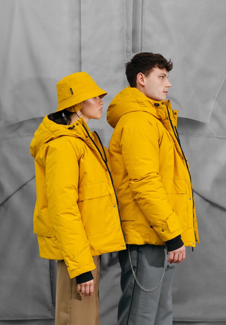 May include: Two people wearing matching yellow hooded jackets and a yellow bucket hat. The jackets have black cuffs and pockets. The background is a light gray color.