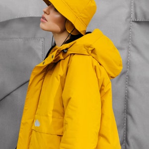 May include: A bright yellow hooded jacket and matching bucket hat. The jacket has a large hood, a front pocket, and a small logo. The hat has a brim and ear coverings. The background is a gray textured surface.