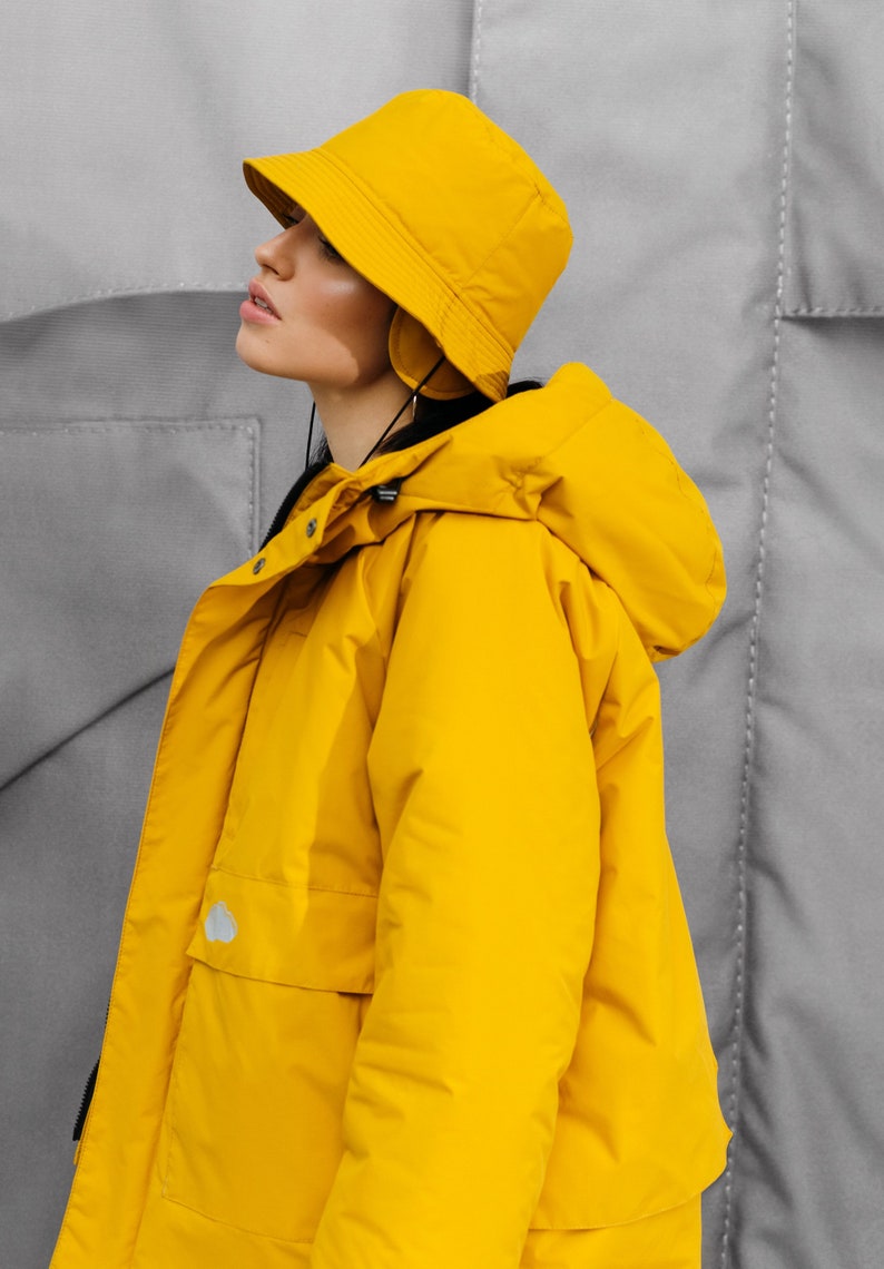 May include: A bright yellow hooded jacket and matching bucket hat. The jacket has a large hood, a front pocket, and a small logo. The hat has a brim and ear coverings. The background is a gray textured surface.