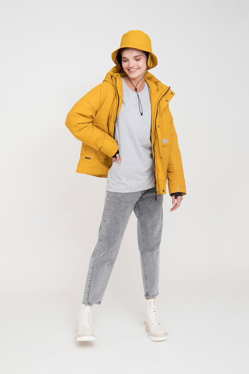 May include: A mustard yellow hooded jacket and matching bucket hat. The jacket has a zip closure and pockets. The outfit includes a gray t-shirt, gray jeans, and white boots. The person is smiling.