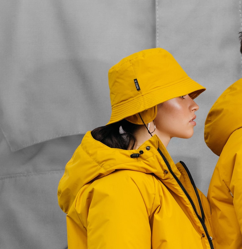 May include: A person wearing a bright yellow bucket hat and matching jacket. The hat has a small black tag on the side. The jacket has a zipper and a hood. The background is a light gray.