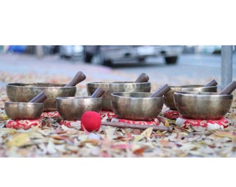 May include: A set of seven silver singing bowls with wooden strikers, arranged on red cushions and surrounded by autumn leaves.