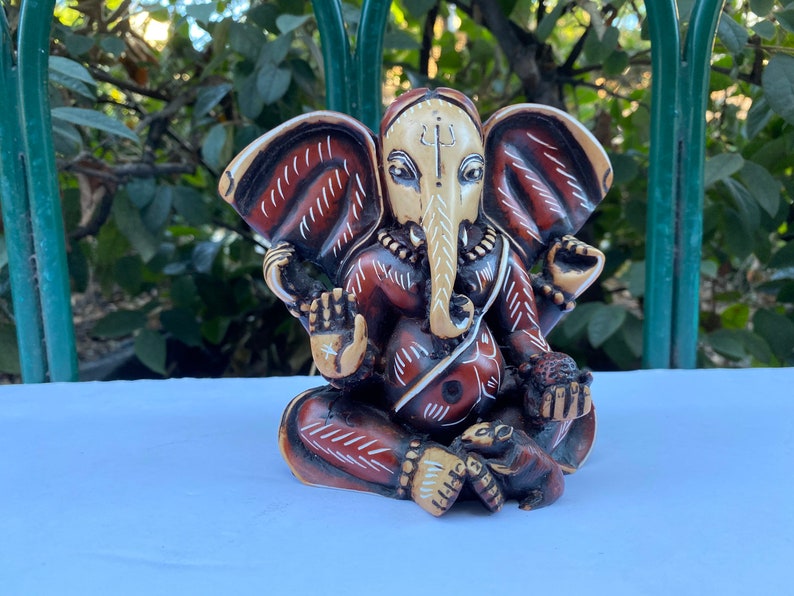 K&ouml;nnte beinhalten: Eine kleine, handbemalte Figur aus Holz des hinduistischen Gottes Ganesha. Die Figur sitzt mit gekreuzten Beinen und erhobenem R&uuml;ssel. Die Figur hat ein rot-wei&szlig;es Muster.
