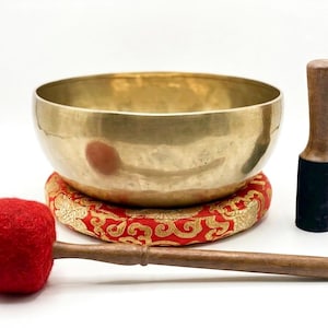 May include: A golden singing bowl with a red and gold patterned cushion, a wooden mallet with a red felt head, and a wooden striker with a black base. The bowl is round and sits on the cushion.