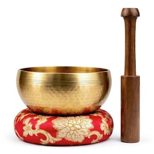 Könnte beinhalten: Eine goldene Klangschale mit strukturierter Oberfläche, die auf einem roten Kissen mit goldenem Blumendesign ruht. Ein Holzschlägel steht neben der Schale. Das Set ist für Meditation und Klangheilung konzipiert. Die Schale hat etwa 15 cm Durchmesser.