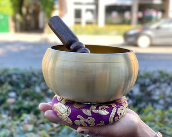 Puede incluir: Un cuenco tibetano de latón con un percutor de madera oscura descansa sobre un cojín morado con bordados dorados. El cuenco es redondo y pulido, reflejando la luz. El cojín es circular y está en la palma de una mano.