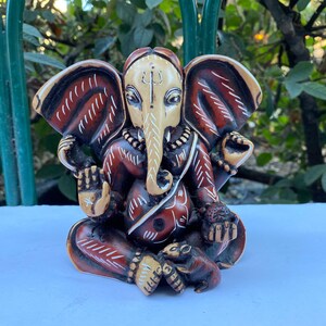 K&ouml;nnte beinhalten: Eine handbemalte Holzstatue von Ganesha, dem hinduistischen Gott der Weisheit und des Wohlstands. Die Statue ist braun und rot mit wei&szlig;en Akzenten und zeigt Ganesha sitzend mit seinem Elefantenkopf und R&uuml;ssel, der ein Bonbon in der Hand h&auml;lt. Eine kleine Maus befindet sich an seinen F&uuml;&szlig;en.
