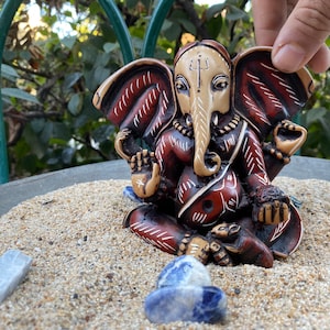 K&ouml;nnte beinhalten: Eine kleine, rot und wei&szlig; bemalte Statue von Ganesha, dem hinduistischen Gott der Anf&auml;nge, des Wohlstands und des Gl&uuml;cks. Die Statue sitzt auf einem Sandbett mit blauen Steinen drumherum.