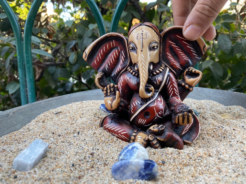 K&ouml;nnte beinhalten: Eine kleine, rot und wei&szlig; bemalte Statue von Ganesha, dem hinduistischen Gott der Anf&auml;nge, des Wohlstands und des Gl&uuml;cks. Die Statue sitzt auf einem Sandbett mit blauen Steinen drumherum.