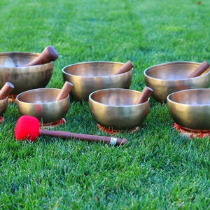 May include: Seven brass singing bowls of different sizes, each with a wooden mallet, are arranged on green grass. The bowls sit on small red and gold patterned cushions. A red mallet lies in the foreground.