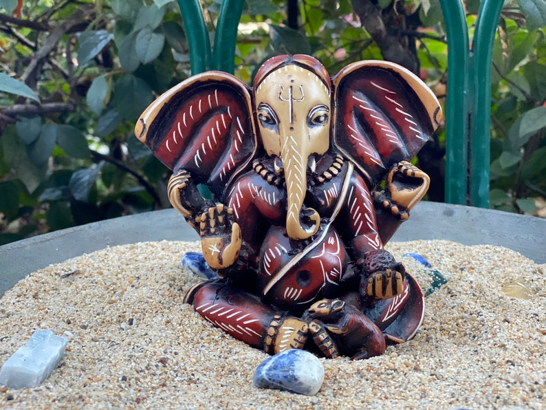 K&ouml;nnte beinhalten: Eine kleine, kunstvoll geschnitzte Statue von Ganesha, dem hinduistischen Gott der Anf&auml;nge, Weisheit und des Wohlstands. Die Statue ist aus Holz und in Rot-, Wei&szlig;- und Braunt&ouml;nen bemalt. Sie steht auf einem Sandbett mit blauen und wei&szlig;en Steinen.