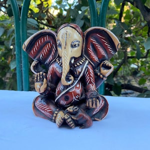 K&ouml;nnte beinhalten: Eine handbemalte Ganesha-Statue in Rot und Wei&szlig; mit filigranen Details. Die Statue sitzt in meditativer Haltung mit eingerolltem R&uuml;ssel.