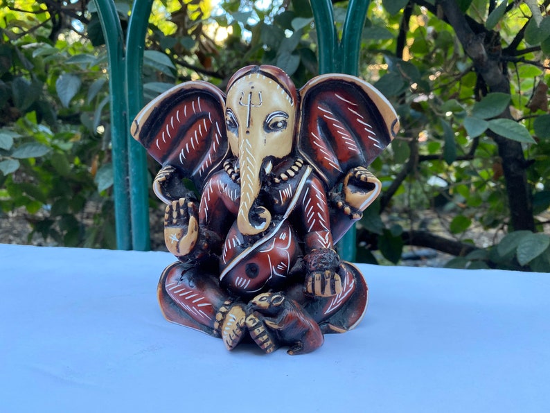 K&ouml;nnte beinhalten: Eine handbemalte Ganesha-Statue in Rot und Wei&szlig; mit filigranen Details. Die Statue sitzt in meditativer Haltung mit eingerolltem R&uuml;ssel.