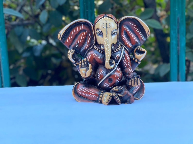 K&ouml;nnte beinhalten: Eine kleine, kunstvoll geschnitzte Statue der hinduistischen Gottheit Ganesha. Die Statue ist aus hellem braunen Material gefertigt und mit roten, wei&szlig;en und schwarzen Akzenten bemalt. Ganesha ist mit einem Elefantenkopf, einem menschlichen K&ouml;rper und vier Armen dargestellt. Er h&auml;lt verschiedene Gegenst&auml;nde in seinen H&auml;nden, darunter eine Lotusblume, ein S&uuml;&szlig;es und ein Buch.