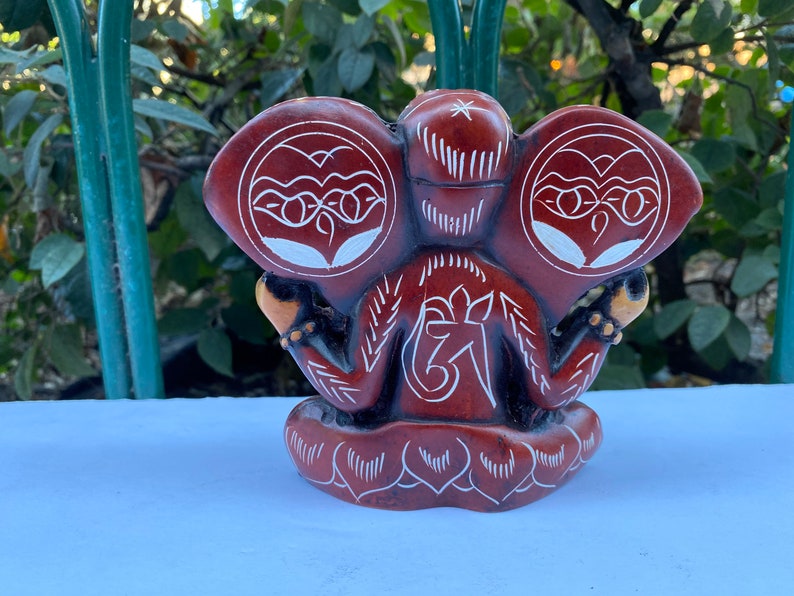 K&ouml;nnte beinhalten: Eine handgeschnitzte Steinskulptur von Ganesha, dem hinduistischen Gott der Weisheit und des Wohlstands. Die Skulptur weist komplizierte Details und ein wei&szlig;es Om-Symbol auf der Vorderseite auf.