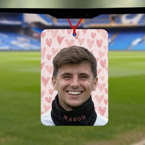 May include: A pink car air freshener with a watercolour design of hearts. The air freshener features a photo of a person with the name "MASON" printed below the photo.