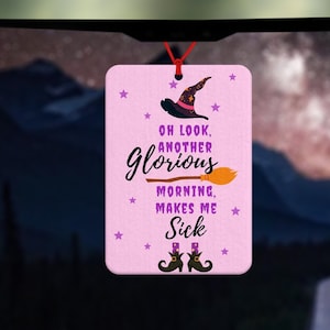 Peut inclure: Un désodorisant de voiture rectangulaire rose avec le texte "Oh look, another glorious morning, makes me sick." Le design comprend un chapeau de sorcière, un balai et des bottes. Suspendu par une ficelle rouge.
