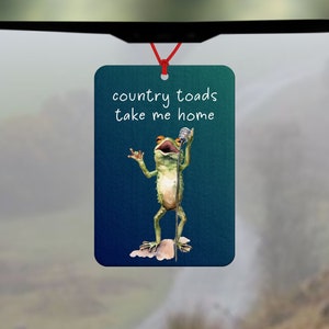 Pode incluir: Um aromatizador de carro azul com um sapo verde cantando em um microfone. O texto no aromatizador diz "country toads take me home".