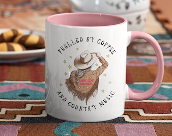 Puede incluir: Taza de cerámica blanca con borde y asa de color rosa. La taza presenta un gráfico de una mujer con un sombrero de vaquero y una chaqueta con el texto "Long Live Cowgirls". El texto "Fueled by coffee and country music" rodea el gráfico.
