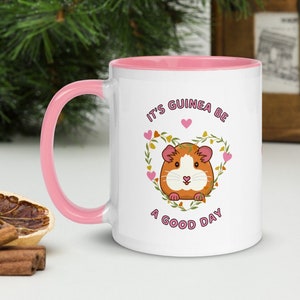 May include: White ceramic mug with a pink interior and handle. The mug features a cartoon guinea pig inside a floral heart, with the text "It's Guinea Be A Good Day" in pink.