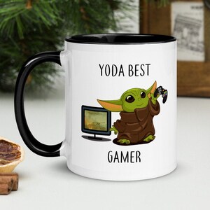 Op de afbeelding: Witte keramische mok met een zwarte rand en handvat. De mok heeft een cartoon afbeelding van een groene baby Yoda die een videogamecontroller vasthoudt en voor een televisie staat. De tekst "YODA BEST GAMER" is op de mok gedrukt.