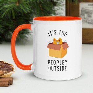 May include: White ceramic mug with an orange rim and handle. The mug has a graphic of an orange cat in a box with the text "It's too peopley outside".