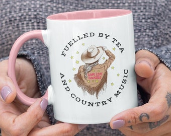 May include: White ceramic mug with a pink rim and handle. The mug features a graphic of a woman wearing a cowboy hat and a denim jacket with the text "Love Live Country" on the back. The text around the graphic reads "Fuelled by Tea and Country Music".