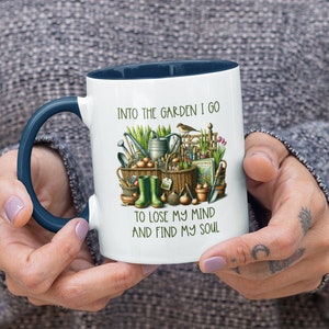 May include: White ceramic mug with a blue rim and handle. The mug features a colorful illustration of gardening tools and the text "Into the garden I go to lose my mind and find my soul."