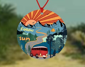 May include: A round car air freshener with a retro design. It features a red van driving on a winding road through a mountain landscape with a sunset in the background. The text "Follow the sun" is printed on the air freshener.