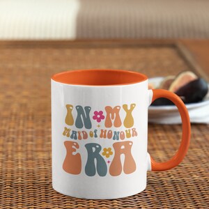 May include: White ceramic mug with an orange rim and handle. The mug has a retro design with the text "In My Maid of Honour Era" in colorful letters.