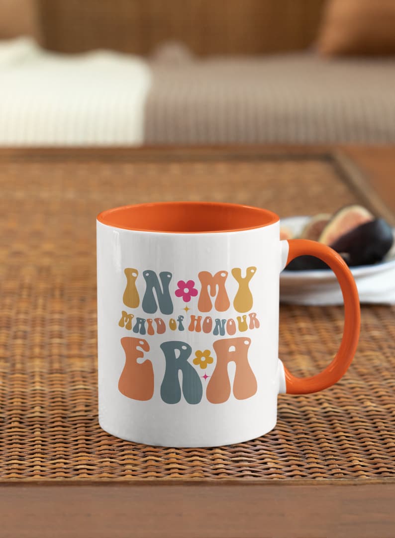 May include: White ceramic mug with an orange rim and handle. The mug has a retro design with the text "In My Maid of Honour Era" in colorful letters.