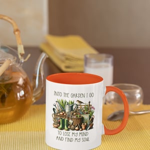 May include: White ceramic mug with an orange rim and handle. The mug features a colorful illustration of gardening tools, plants, and a bird. The text "Into the garden I go to lose my mind and find my soul" is printed on the mug.