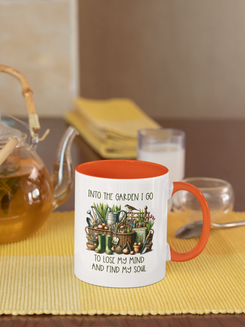 May include: White ceramic mug with an orange rim and handle. The mug features a colorful illustration of gardening tools, plants, and a bird. The text "Into the garden I go to lose my mind and find my soul" is printed on the mug.