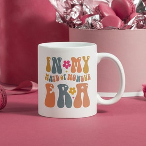 May include: White ceramic mug with a colorful graphic that says "In My Maid of Honour Era" with flowers and stars.