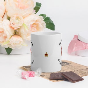 May include: White ceramic mug with black text that reads "You're back, da da da..." and a crown graphic. The mug is sitting on a table with a pink candy, chocolate bars, and a newspaper.