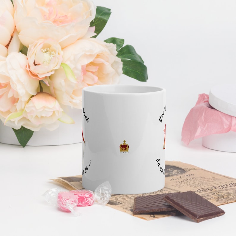 May include: White ceramic mug with black text that reads "You're back, da da da..." and a crown graphic. The mug is sitting on a table with a pink candy, chocolate bars, and a newspaper.