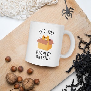 May include: White ceramic mug with a black and orange illustration of a cat in a box. The text "It's too peopley outside" is printed on the mug.